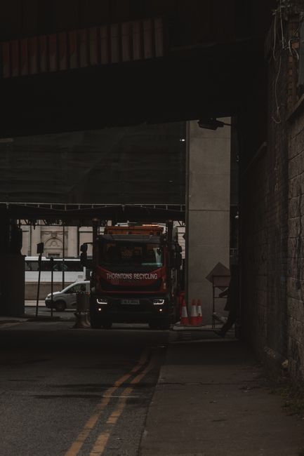 A side view of a large commercial rubbish collection truck parked on an urban street with a backdrop of tall, multicoloured residential buildings and a few trees. The truck features a white cab with a