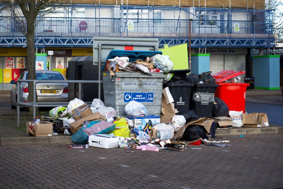 A large pile of mixed domestic rubbish is overflowing from several different waste containers situated on a paved pavement near a street. The pile includes various cardboard boxes, plastic bags, paper