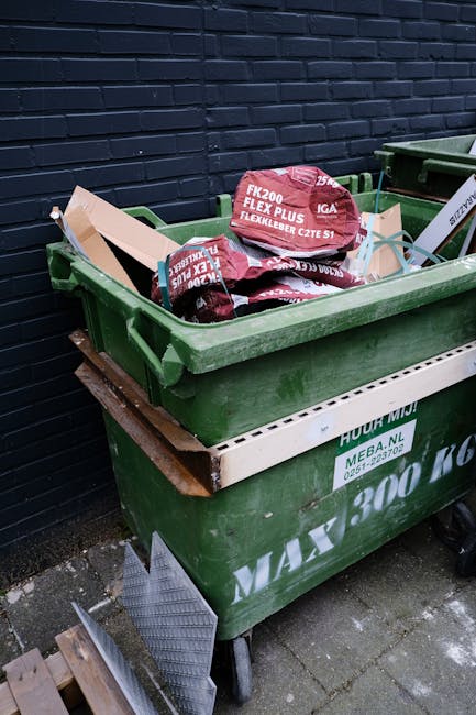 A large pile of mixed domestic rubbish is overflowing from several different waste containers situated on a paved pavement near a street. The pile includes various cardboard boxes, plastic bags, paper